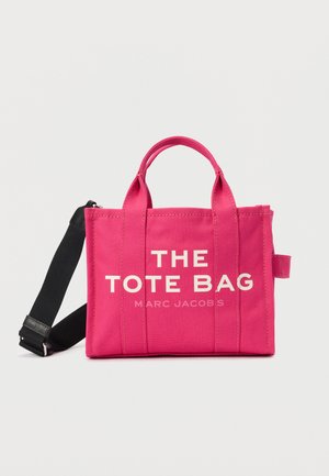 Sac fourre-tout en toile rose avec deux poignées supérieures et une bandoulière noire amovible. Présente un texte blanc audacieux : "THE TOTE BAG" et "MARC JACOBS".