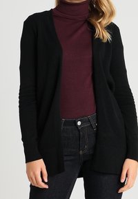 Černý cardigan s texturovaným pletením, design s otevřeným předním dílem, vrstvený přes burgunský rolák. Kombinovaný s tmavými džíny, vytváří neformální vzhled.