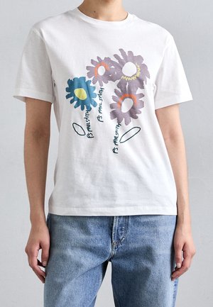 Hvid t-shirt med korte ærmer med fire store abstrakte blomster i blå og lilla, båret med lyseblå jeans på en person, der står mod en ensfarvet baggrund.