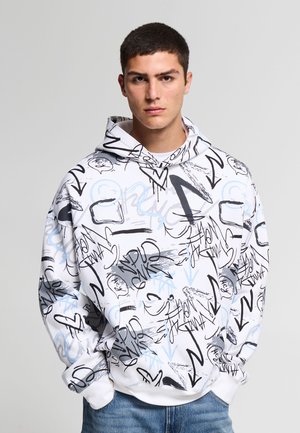 Giovane uomo che indossa una felpa con cappuccio bianca con disegni astratti in stile graffiti neri e blu, abbinata a jeans blu, in piedi davanti a uno sfondo neutro.