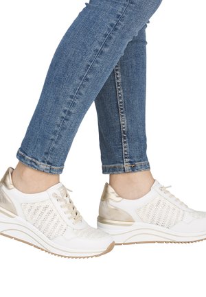 Person iført blå skinny jeans og hvide sneakers med gyldne accenter, stående med den ene fod let hævet.