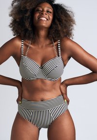 Usměvavá žena s afro účesem, oblečená v černobílém pruhovaném bikinovém topu a horních dílech plavek s vysokým pasem, s rukama opřenýma o boky.