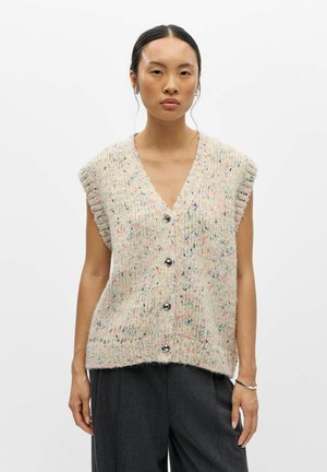 Femme portant un gilet en maille crème sans manches, boutonné, avec des taches multicolores et un pantalon ample gris foncé, debout devant un fond uni.