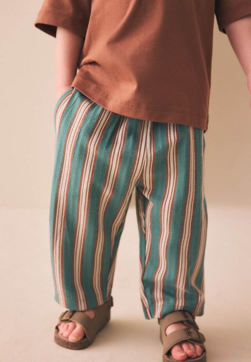 Niño con camiseta marrón, pantalones a rayas turquesa y marrones, con sandalias marrones de punta abierta, de pie sobre un suelo y pared color beige claro.