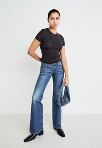 Sort tætsiddende t-shirt, blå flagrende jeans, holder en denimjakke. Glat tekstur, lysvasket denim, rektangulær skæring, sorte spidse støvler.