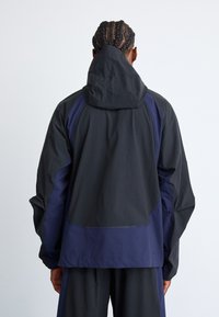 Black and navy windbreaker jacket with a hood, featuring a textured fabric, loose fit, and contrasting side panels.
