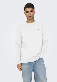 Felpa bianca a girocollo con polsini e orlo a costine. Presenta una piccola toppa con logo rosso sul petto in alto a sinistra. I jeans in denim sono leggermente sbiaditi.