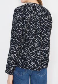 Femme portant un chemisier bleu marine à manches longues avec un motif abstrait beige et un jean bleu, vue de dos sur un fond uni.