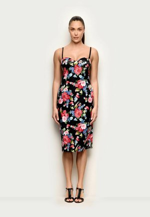 Kobieta stojąca przodem, ubrana w czarną, kwiecistą sukienkę midi na cienkich ramiączkach oraz czarne sandały na wysokim obcasie, na jednolitym tle.