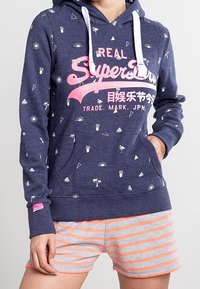 Felpa navy con stampa grafica bianca, featuring palme e motivi solari, e un logo rosa. Abbinata a pantaloni corti a righe grigie e arancioni.