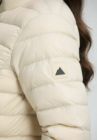 Veste matelassée crème avec des sections en diamant, une texture lisse et un patch logo triangulaire noir contrastant sur la manche.