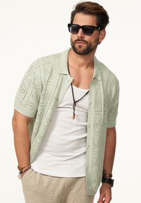 Helles, grünes kurzärmeliges Hemd mit Knopfleiste und offenem Muster, getragen über einem weißen Tanktop. Zubehör umfasst ein dunkles Perlenarmband und Sonnenbrille.