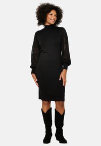 Abito in maglia nera con scollatura alta, silhouette aderente e lunghe maniche in tessuto trasparente a fantasia. Abbinato a stivali neri in suede al ginocchio.
