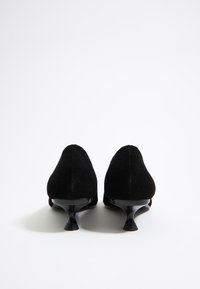 Zapatos de tacón alto de ante negro con puntera afilada y tacones curvados distintivos, que presentan una textura suave y una silueta elegante.
