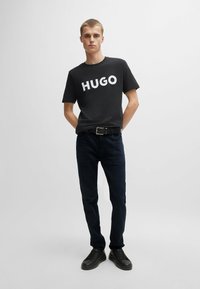 Camiseta negra con estampado blanco "HUGO", combinada con jeans oscuros y zapatillas negras. El modelo se encuentra de pie con las manos en los bolsillos frente a un fondo liso.