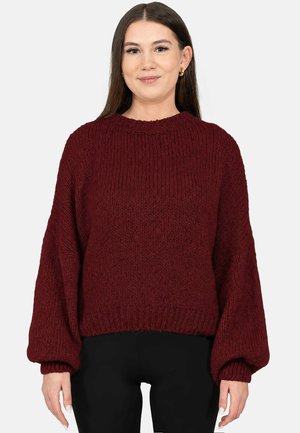 Jeune femme aux longs cheveux bruns portant des boucles d'oreilles en anneaux dorés et un pull en maille bordeaux ample avec des manches ballon ainsi qu'un pantalon noir.