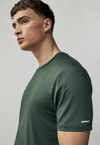 Skovgrøn kortærmet sportstrøje, tekstureret stof med et subtilt mønster, rund halsudskæring og reflekterende detaljer på ærmet.