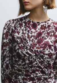 Vestido con estampado floral y mangas largas, con tonos burdeos y morados sobre una tela blanca texturizada. Incluye un pendiente decorativo.