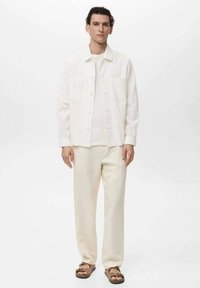 Light-colored linen jacket with two front pockets, paired with a matching shirt and trousers. Sandals complete the outfit.