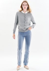 Cardigan grigio con bottoni, maniche lunghe, indossato sopra una camicia bianca con colletto, abbinato a jeans slim-fit blu chiaro e scarpe beige chiare.