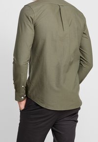 Koszula z długim rękawem w oliwkowej zieleni, wykonana z bawełny, z plisą z tyłu, zaokrąglonym dołem oraz mankietami na guziki z srebrnymi akcentami. Noszona z ciemnymi spodniami.