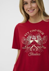 Raudonas medvilninis marškinėliai su aukso spalvos grafiniu dizainu, vaizduojančiu du paukščius, gėlių akcentais ir tekstu "Skristi per aukštai laisvei". Trumpomis rankovėmis ir apvaliu kaklu.