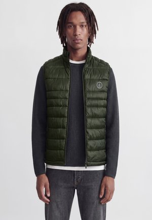 Un modèle porte un gilet matelassé sans manches vert foncé sur un pull gris anthracite et un jean foncé, devant un fond clair et uni.