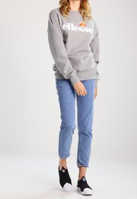 Sweat-shirt gris avec le logo "ellesse", jean bleu denim, et chaussures noires à enfiler avec bouts blancs. Tenue simple et décontractée.