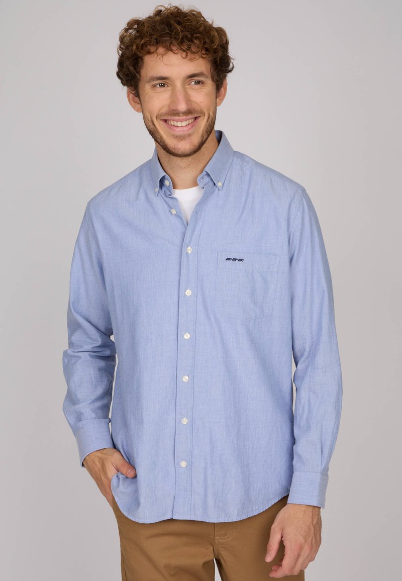 Chemise bleu clair à manches longues en tissu lisse, dotée d'une poche poitrine avec un petit logo et d'un ourlet arrondi.