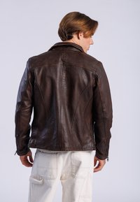 Veste en cuir marron avec un design ajusté, dotée d'un col pointu et de poignets zippés épurés. Coutures notables à l'arrière.