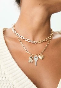 Collane a strati color oro con grandi anelli a catena, dotate di un charm a forma di orsetto, una chiave e un pendente a forma di cuore in varie finiture.