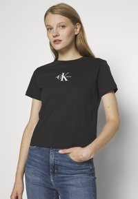 Zwarte katoenen t-shirt met een ronde hals, korte mouwen, en een wit logo op de borst. Combinatie met blauwe denim jeans.