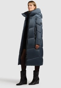 Dlhý tmavomodrý prešívaný kabát s vysokým golierom, zapínaním na zips a geometrickým prešívaným vzorom. Má bočné vrecká a voľný strih.