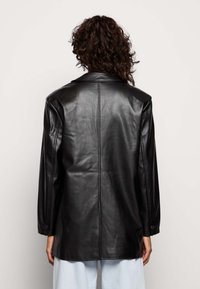 Henkilö, jolla on kihara hius, pukeutuneena mustaan nahkatakkiin, näkyy takaa, vaaleat housut päällään yksinkertaisen taustan edessä.