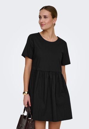 Vrouw draagt een korte zwarte jurk met korte mouwen, houdt een donkerbruine handtas vast en draagt een gouden armband aan haar linkerpols.