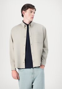 Camicia button-up beige con taschino sul petto, realizzata in tessuto testurizzato. Indossata sopra un top scuro, abbinata a jeans azzurri chiari.