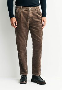 Brown corduroy pants with vertical ribbing, pleats at the waist, and a button closure. Paired with black shoes, featuring a textured surface.