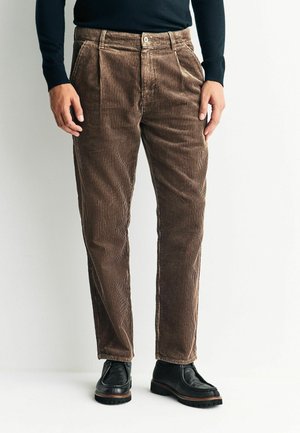 Brown corduroy pants with vertical ribbing, pleats at the waist, and a button closure. Paired with black shoes, featuring a textured surface.