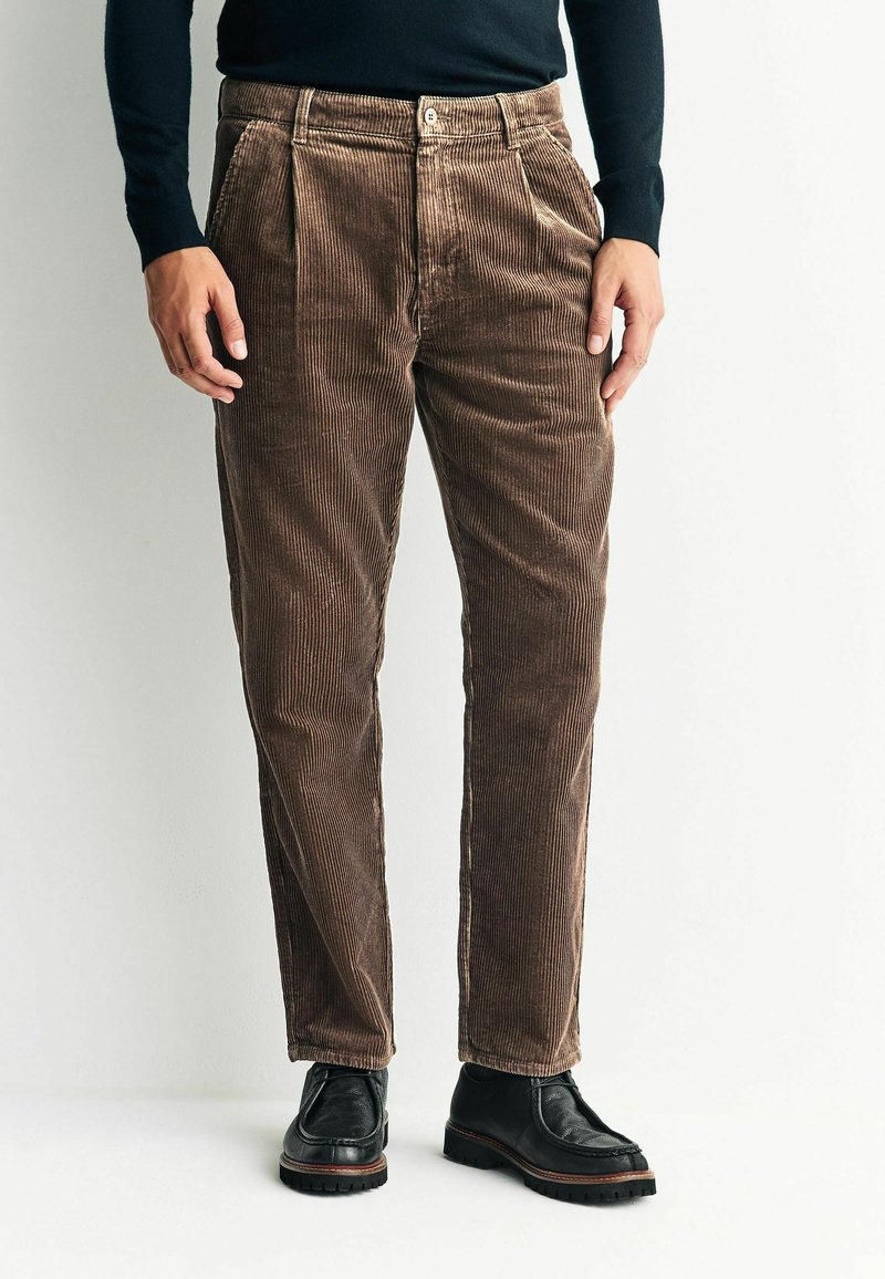 Brown corduroy pants with vertical ribbing, pleats at the waist, and a button closure. Paired with black shoes, featuring a textured surface.