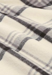 Pătură din fleece de culoare crem, cu un model în carouri negre și gri. Textura moale și pliurile în strat creează adâncime și interes vizual.