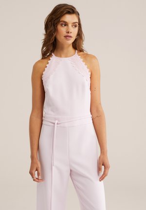 Nő, aki ujjatlan, világos rózsaszín overált visel, csipkedíszítéssel a vállakon és vékony övvel, semleges háttér előtt áll.