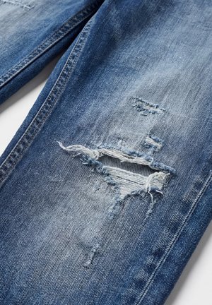 Plave traperice s horizontalnim razderotinom i izblijedjelim nitima na području bedra, prikazujući istrošenu teksturu tkanine i detalje šava.