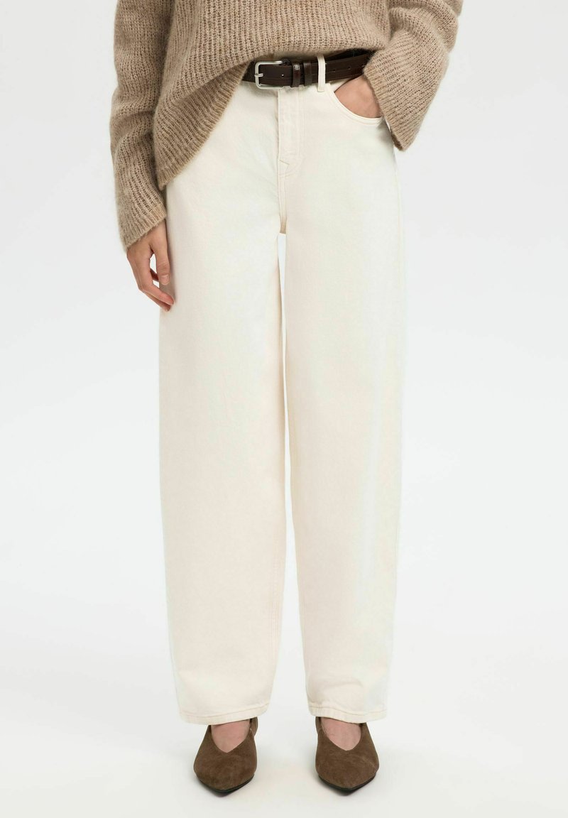 Personne portant un pantalon large crème, une ceinture marron, des chaussures en daim marron à bouts pointus, et un pull en tricot beige ample avec une main dans la poche.