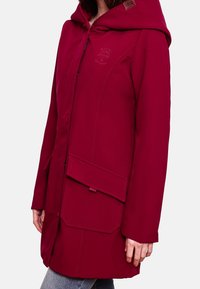 Veste imperméable cramoisie avec capuche, dotée d'un zip sur le devant, d'une grande poche avant et d'un logo discret sur la poitrine. Texture douce.