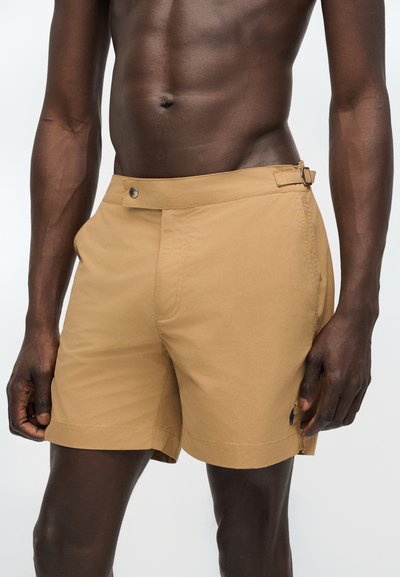 Ingek nélküli férfi bézs, oldalsó állító csattal ellátott, szabott rövidnadrágban, világos, egyszínű háttér előtt áll.