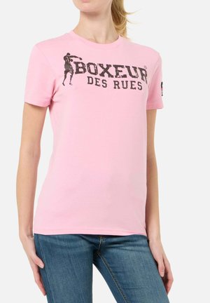 Femme portant un t-shirt rose avec le texte "BOXEUR DES RUES" et la silhouette d'un boxeur, associé à un jean bleu sur un fond uni.