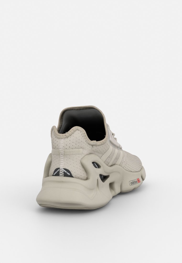 Zapatilla deportiva beige con un upper de malla texturizada, interior negro y suela esculpida distintiva. Presenta tres acentos a rayas en el lateral.