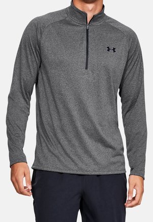 Mann trägt einen grauen Pullover mit langen Ärmeln und einem Viertel-Reißverschluss mit kleinem schwarzem Under Armour-Logo auf der linken Brust.