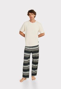 Kurzärmliges cremfarbiges Baumwoll-T-Shirt, kombiniert mit lockeren schwarzen Hosen, die ein weißes und grünes geometrisches Muster aufweisen.