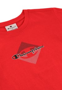 Czerwona bawełniana koszulka z okrągłym dekoltem, z dużym logo w postaci diamentu z szaro-czarnymi paskami i napisem "Champion" w centrum.